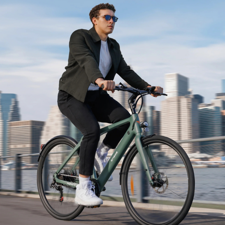 a man wearing sunglasses, wearing black jeans and t-shirt, white trainers. enjoying a a lovely summers day on his Green Engwe N1 Air Carbon fiber e-bike along the river with city in background