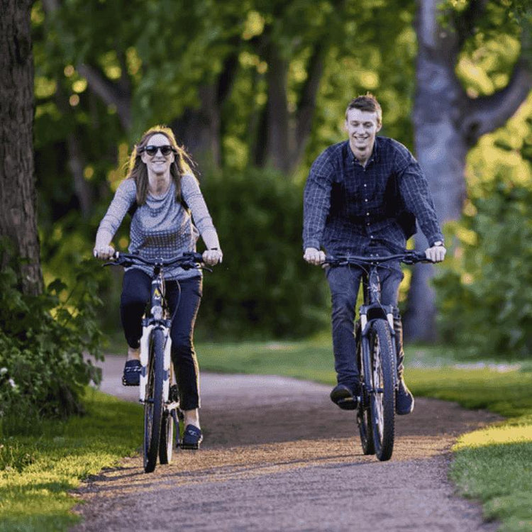 A couple riding towards you next to each other, both wearing grey and black on a cycle path in a forest with sun shining through trees