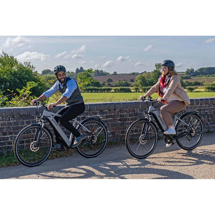 A man with black trousers, blue shirt and black waist coat and lady wearing cream with red scarf both of them with black helmets riding grey bikes next to old wall with county scenery the other side on a sunny day