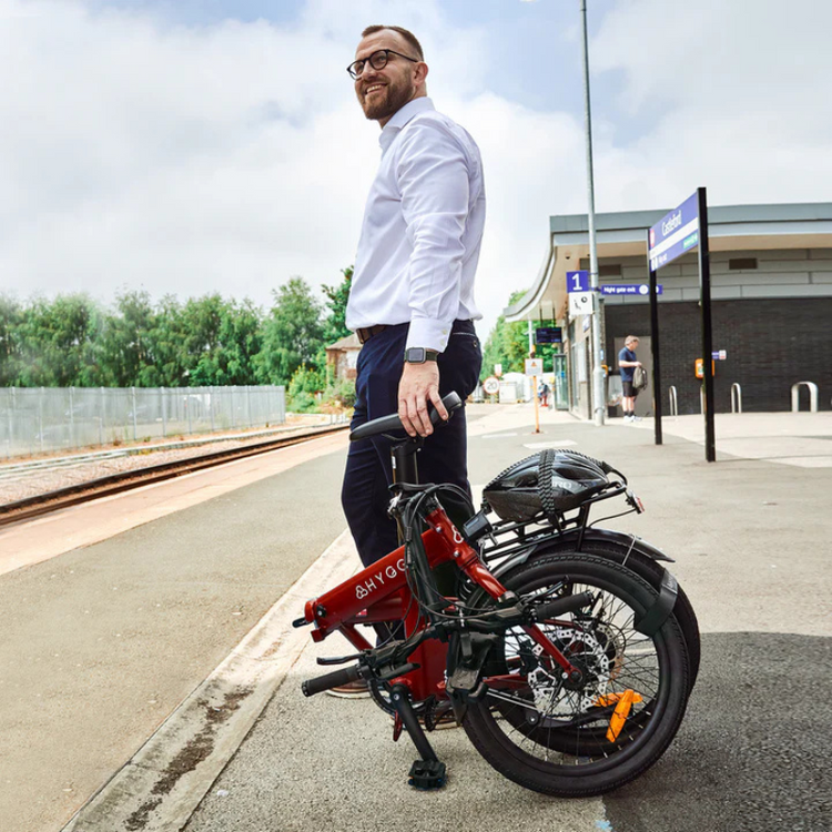 Man with beard in white shirt and trousers standing next to folded red Hygge Hygge bike on train station platform on sunny day