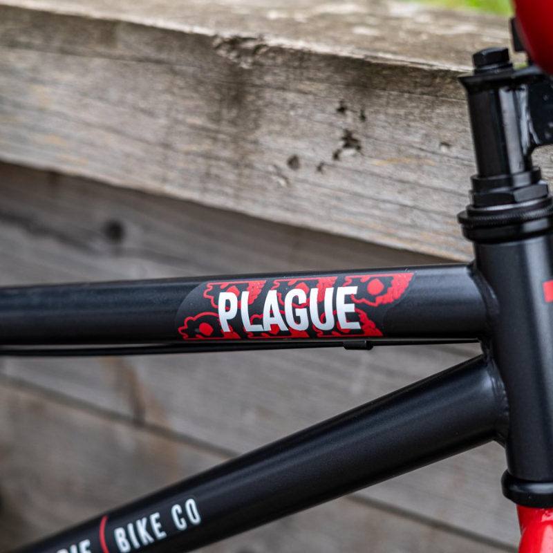 Close-up of a bicycle frame with 'Plague' and 'Bike Co' branding against a wooden background.