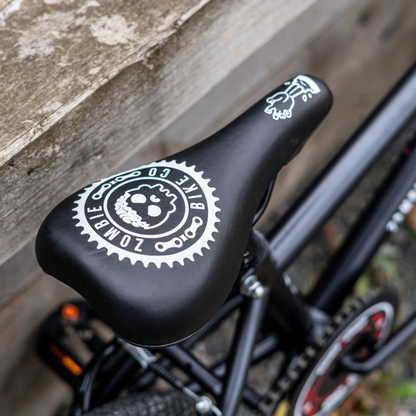 Bicycle seat with a Zombie Bike Co logo leaning on a wooden fence